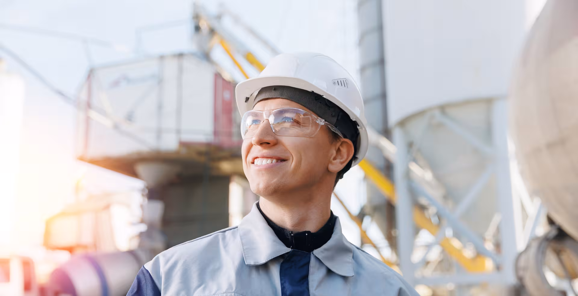 Technicien de chantier portant un casque et des lunettes de protection sur un site industriel, regard tourné vers l’horizon.