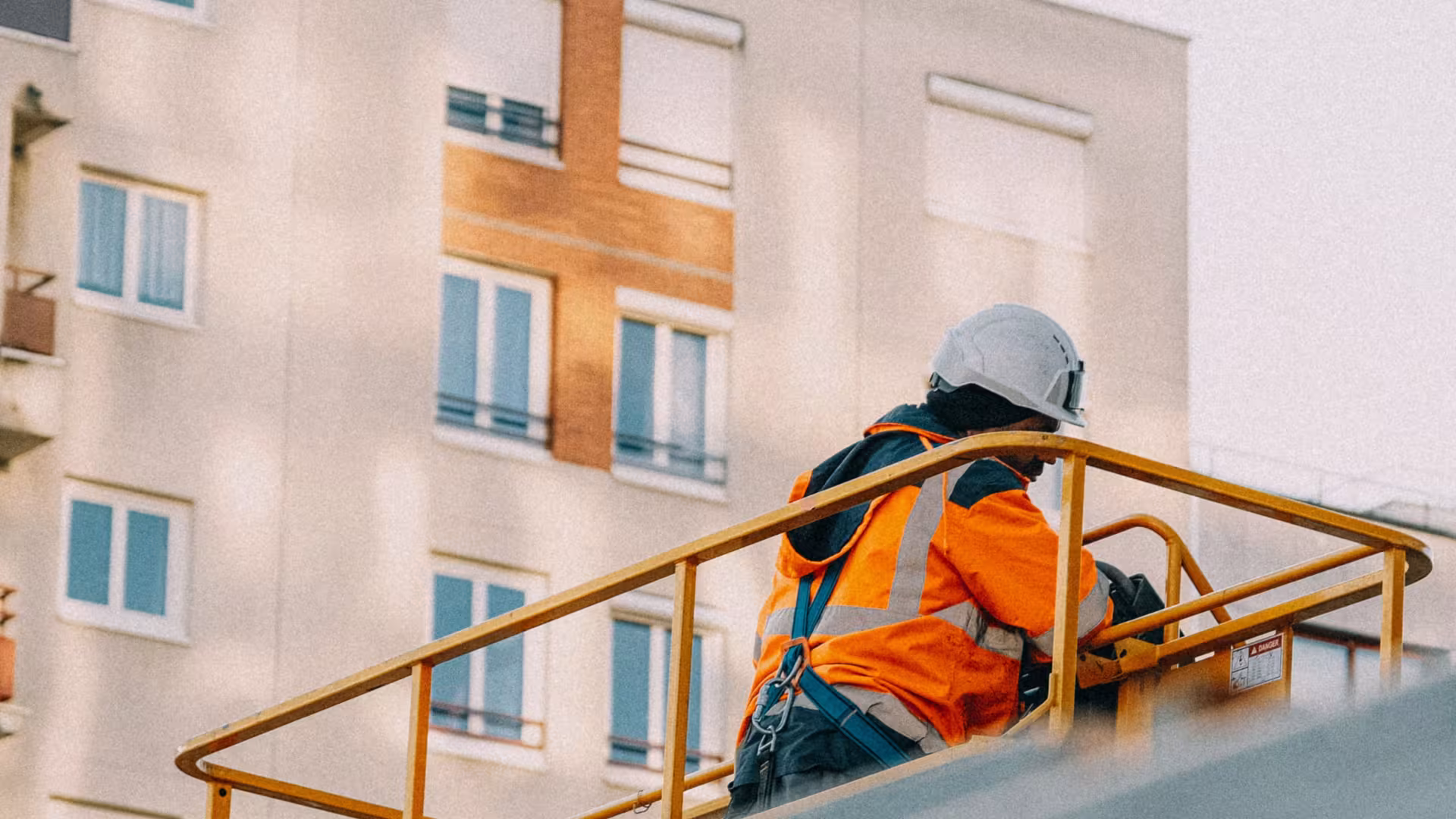 Ouvrier équipé d’un casque, d’un harnais et d’un gilet haute visibilité intervenant en hauteur devant un immeuble résidentiel.